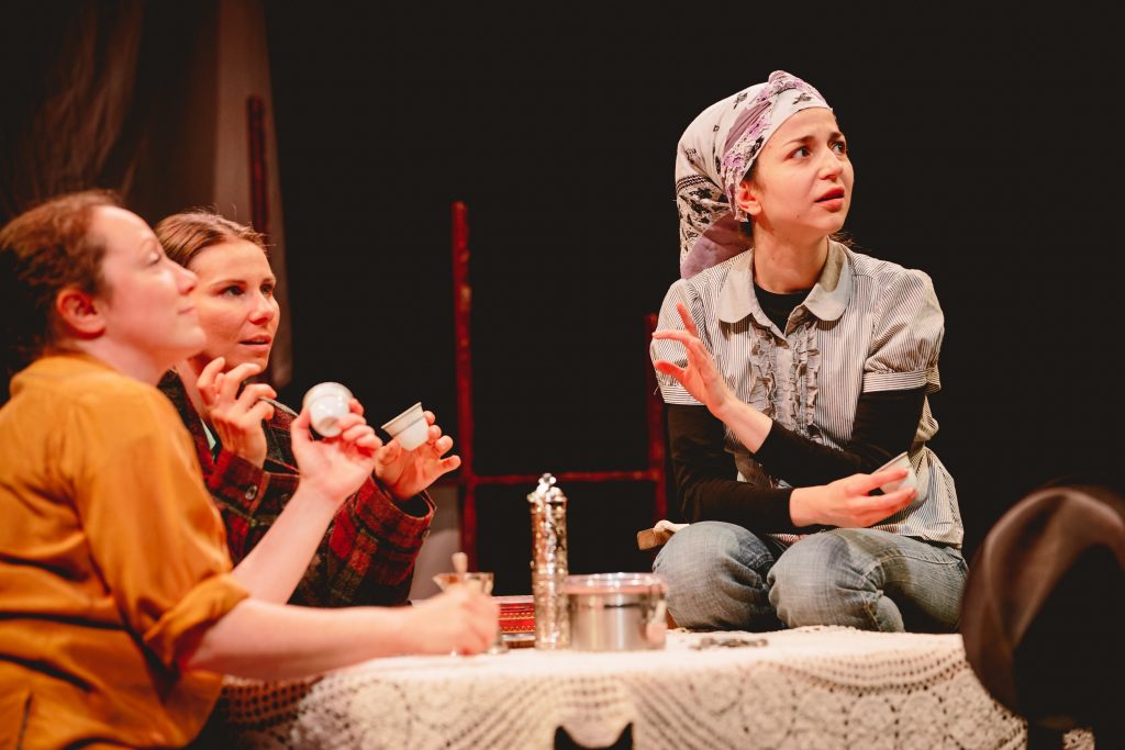 Three female actors sit around, and on, a table covered in a lace tablecloth, examining the contents of their coffee cups.