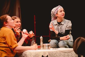 Three female actors sit around, and on, a table covered in a lace tablecloth, examining the contents of their coffee cups.