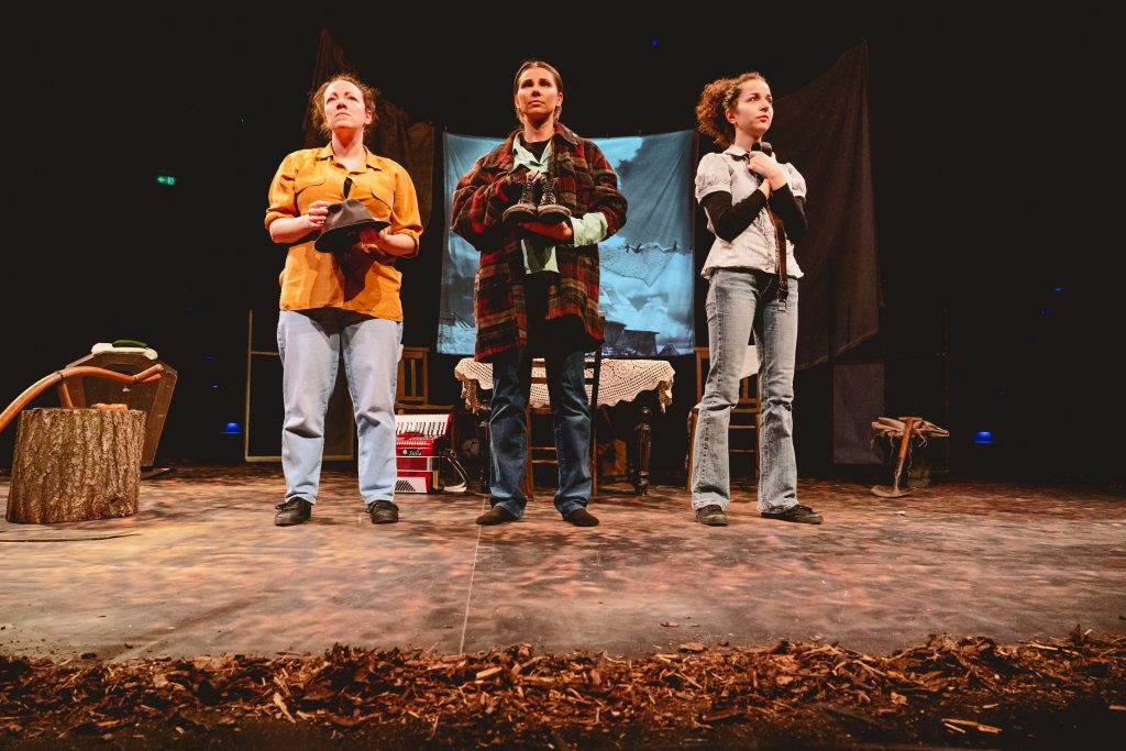 Three female actors stand in a row holding a hat, some shoes, and a belt respectively, and look into the distance.