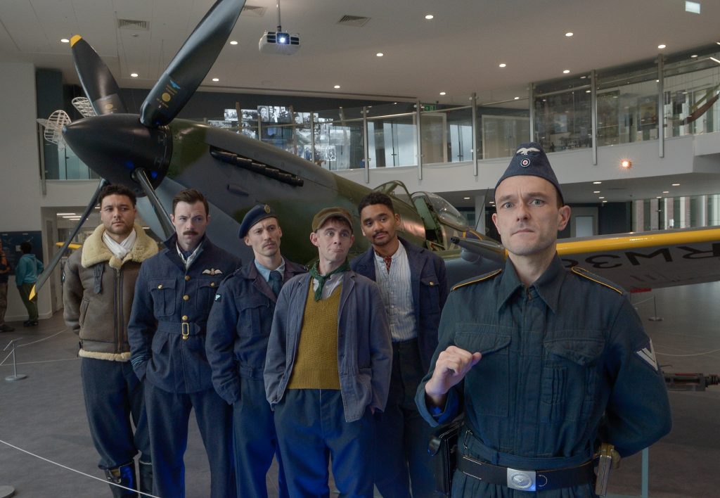 Five actors playing prisoners of war of multiple nationalities in a variety of costumes stand next to a Spitfire plane at the Potteries Museum & Art Gallery looking over at an actor in a Luftwaffe costume in the foreground.