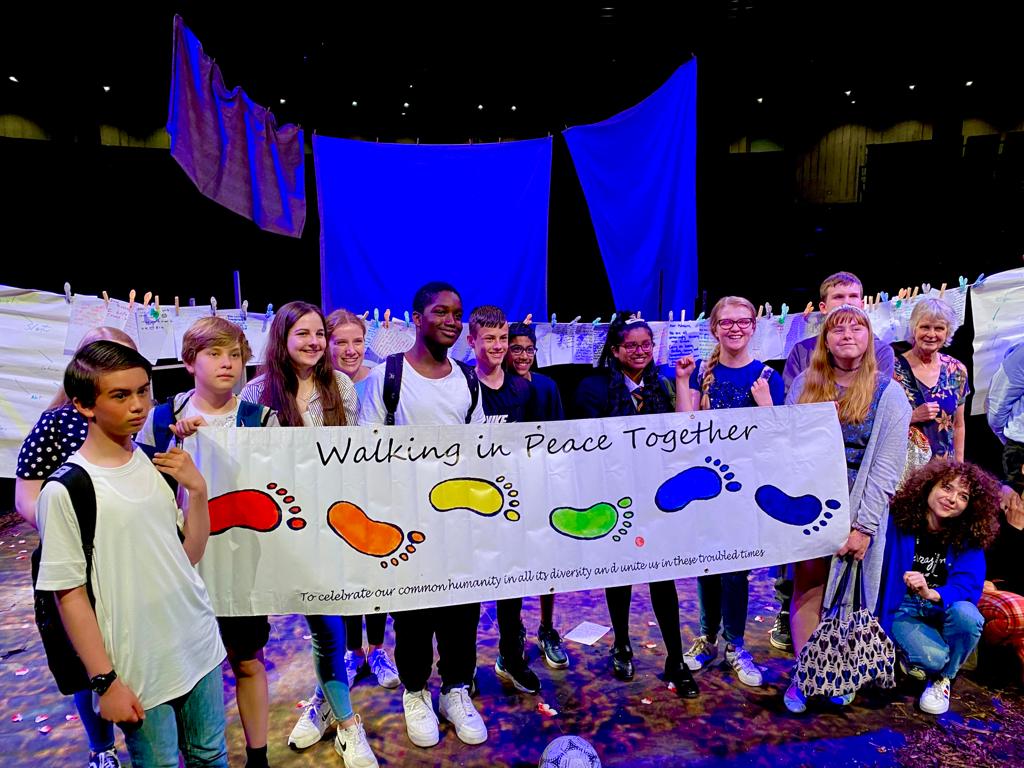 A group of young people stand holding a banner which has different coloured footprints and says ' Walking In Peace Together'