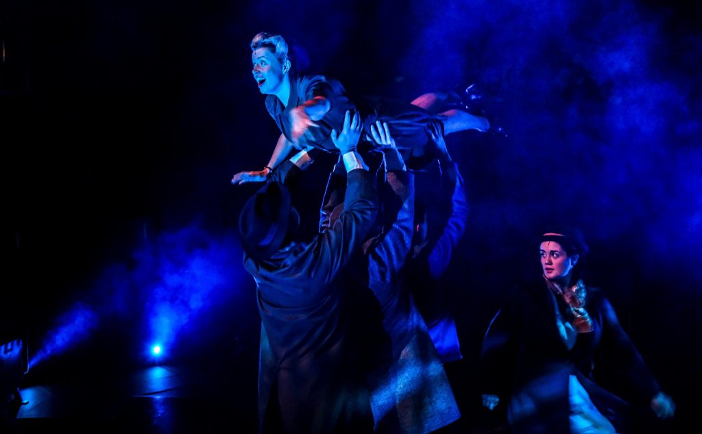 Three shadowy figures in coats and jackets lift a female actor dressed in 1930s clothing into the air, surrounded by smoke. There is another female actor who looks over at them.