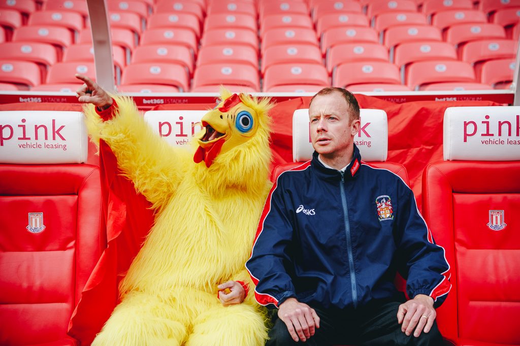 An actor (Michael Hugo) playing Neil Baldwin dressed as a chicken sits with another actor (Gareth Cassidy) as Lou Macari, sat in the dugout at Stoke City FC's bet365 stadium.   