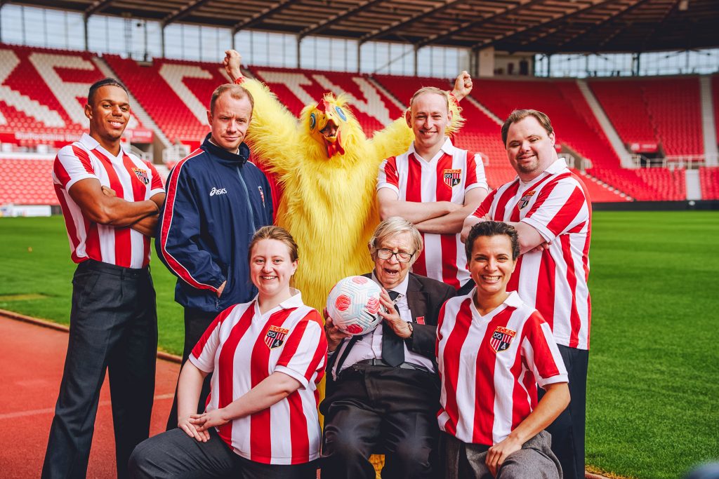 Five actors in Stoke City Football Club kits pose pitchside with two actors dressed as former Stoke City FC manager Lou Macari and Neil Baldwin dressed as a chicken. The real Neil Baldwin sits with them holding a football.  