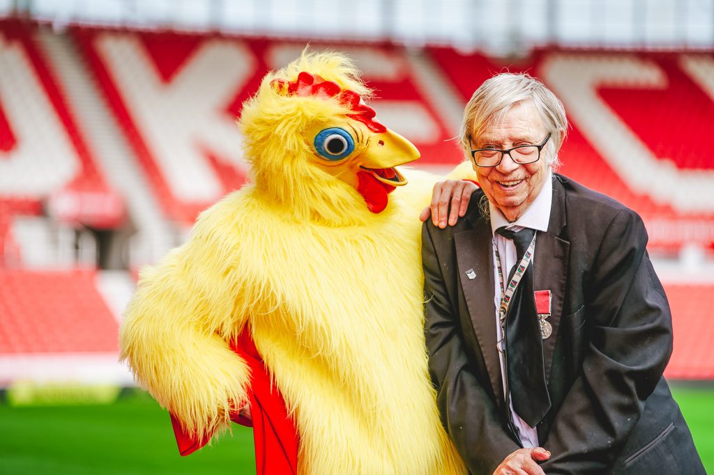 An actor (Michael Hugo) playing former Stoke City kitman Neil Baldwin dressed as a chicken leans on the real Neil Baldwin as they laugh together at Stoke City FC's bet365 stadium. 