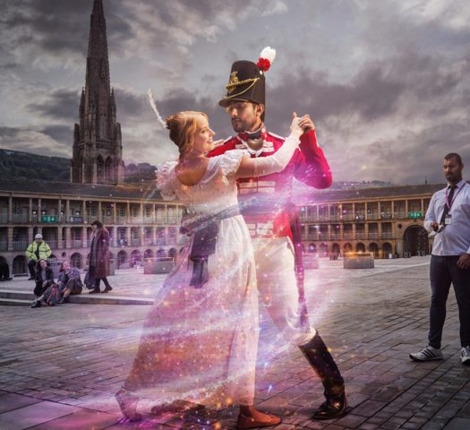 Two people in regency dress dance on a city square as people in modern dress look on.