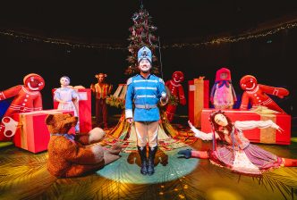 The Nutcracker stands in front of an ornate Christmas tree surrounded by toys including a teddy bear, china doll, rag doll, gingerbreads, cymbal monkey and a nesting doll. Production picture from The Nutcracker at the New Vic. Credit Andrew Billington.