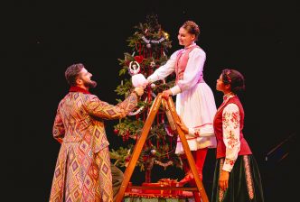 A family decorate a Christmas tree. Marie, a child, stands on a ladder and is passed the Sugar Plum Fairy decoration. Production picture from The Nutcracker at the New Vic. Credit Andrew Billington.