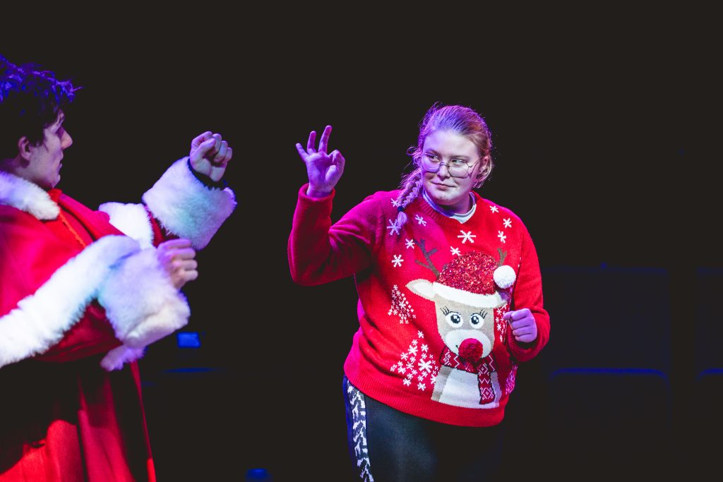 A girl makes an 'okay' gesture with her hands at a boy in a Father Christmas outfit. Young people dance in celebration. Picture from the Borderlines Young People's Theatre Company's version of The Nutcracker 2023. Credit: Andrew Billington