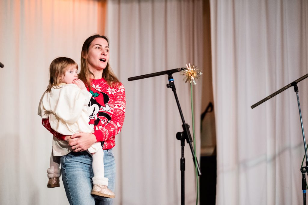A woman sings into a microphone holding a toddler in her arms. Picture from the Vertep at Newcastle Congregational Church, performed by Ukrainian families. Credit: Jenny Harper. 