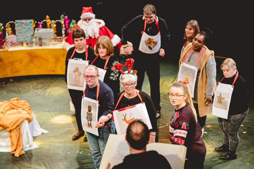 A group of people gather, one in a Father Christmas outfit, one in a Christmas jumper, and the others wear pictures of Christmas-related items, decorations and presents round their necks. Picture from the Strathcross version of The Nutcracker 2023. Credit: Andrew Billington