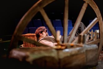 Moshe is resting, his head on a wicker basket. He is holding a letter. He's seen through a wooden wheel in the foreground. Credit: Andrew Billington