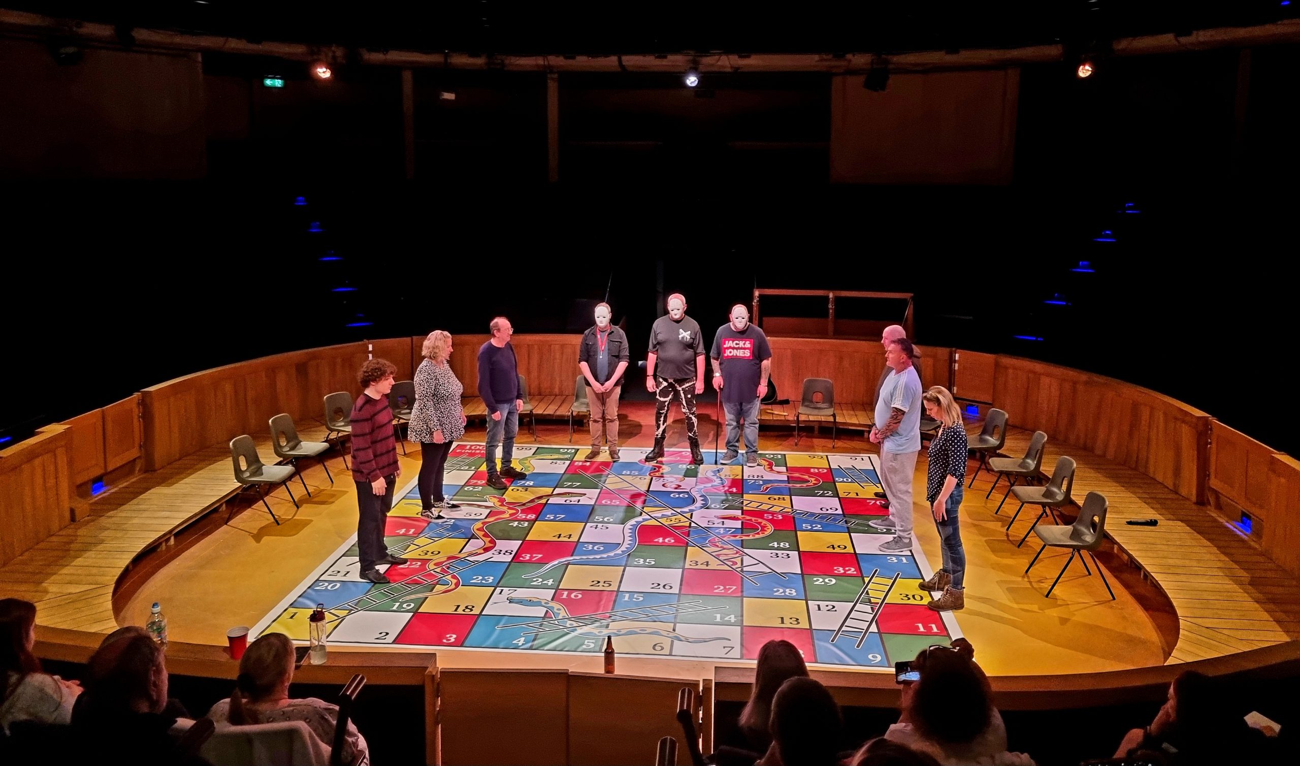The Next Chapter drama group perform on the New Vic Theatre stage for World Mental Health Day 2023. They are stood along the edge of a snakes and ladders games board, three of the performers facing the audience wear masks.