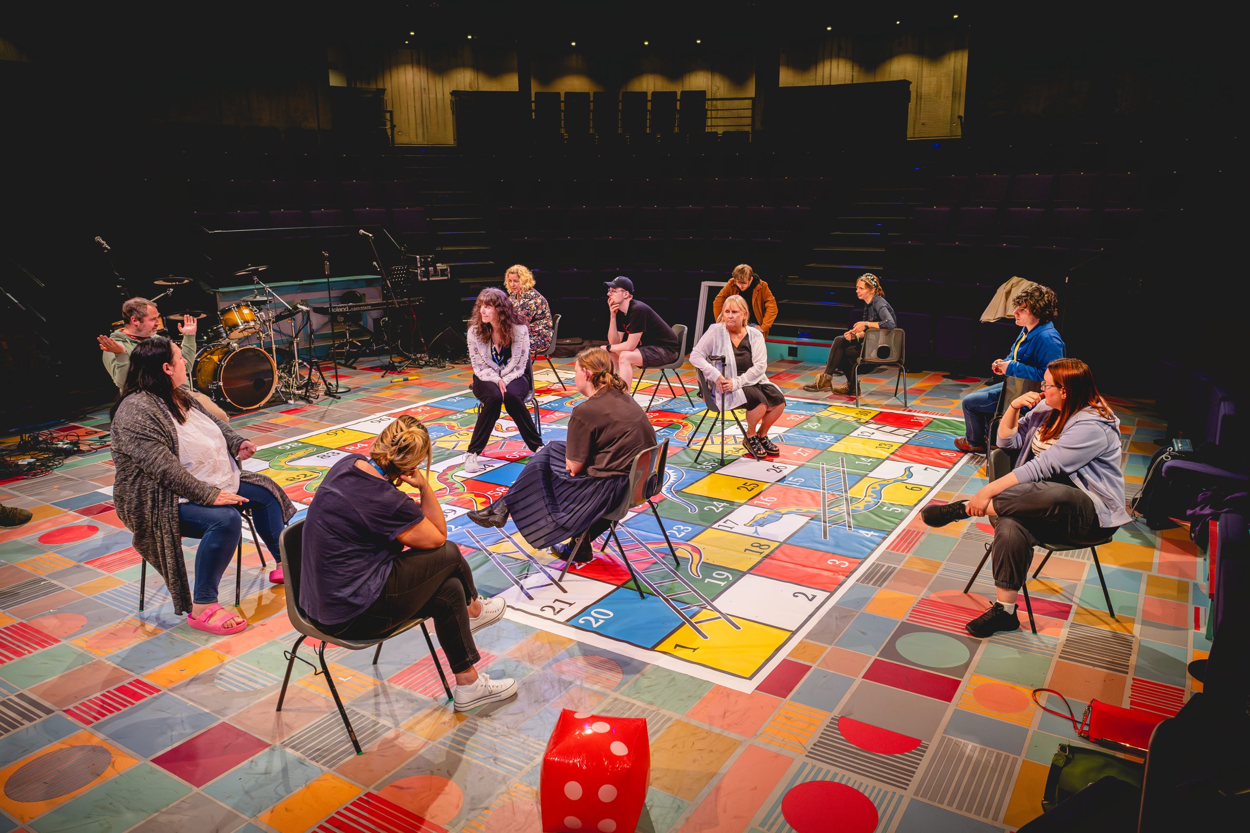 New Vic Borderlines' Next Chapter group sit on chairs scattered around a giant snakes and ladders board on the New Vic's stage