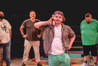 A man in green trousers, a white t-shirt and grey jacket holds a giant pencil up to his ear as if it's a telephone, during a drama game.